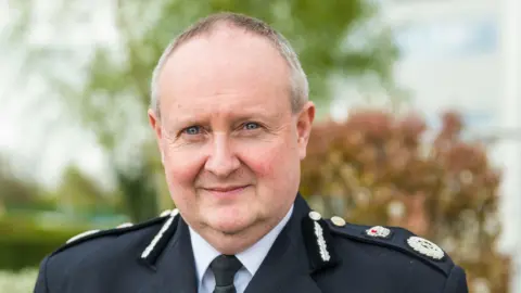 Cheshire Police Chief Constable Mark Roberts in his police uniform. He is a middle-aged man with receding light grey hair and blue eyes. There are trees in the background.