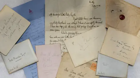 A top-down view of several handwritten letters and envelopes, which are clearly very old. The paper they are written on is of various colours and one of the envelopes is sealed with wax.