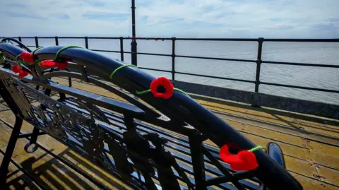 A black iron bench with knitted poppies woven around its frame. The bench is on Southend Pier and faces out to sea.
