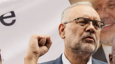 A bespectacled Ebrahim Rasool raises a fist in front of a poster bearing his face