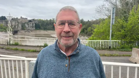 The image shows an older man wearing a grey jumper standing in front of a bridge. Behind him, a river with brown water flows beneath the structure, and an old castle can be seen in the background.
