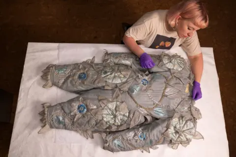 David Parry/PA Media Assignments A grey clown's costume with flower patterns is displayed on a table with a museum curator examining the fabric