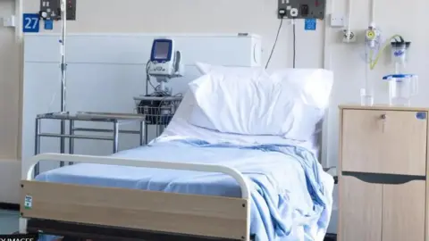 An empty hospital bed with a light blue cover and a rumpled pillow. A bedside cabinet with a jug and glass of water is to the right and some medical equipment to the left