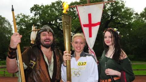 LOCOG via Getty Images JUNE 28: In this handout image provided by LOCOG, Torchbearer 037 Laura Graves stands in front of The Major Oak in Sherwood Forest whilst holding the Olympic Flame alongside 21st century Robin Hood Ade Andrews and Sylvia Robson on Day 41 of the Olympic Flame Torch Relay leg on June 28, 2012 in Nottingham, England. The Olympic Flame is now on day 41 of a 70-day relay involving 8,000 torchbearers covering 8,000 mile