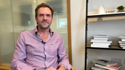Alex Whitmore pictured seated in a wooden chair in an office setting, facing the camera. He is wearing a pink and white striped button-down shirt. The background includes a glass wall with a wooden frame on the left and a black metal shelving unit on the right holding stacked books and a small architectural model.