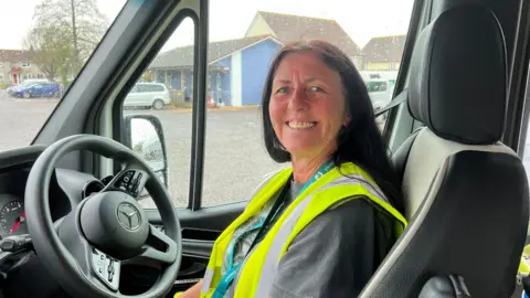 A woman in a high-vis jacket. She is smiling and looking at the camera. She is sitting in the driver's seat of a minibus. In the background there is a bus depot.