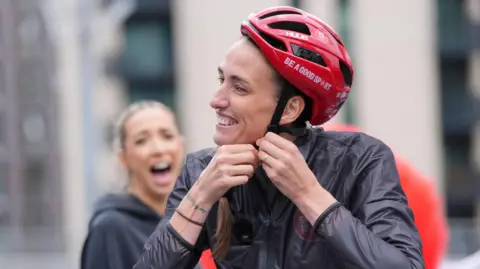 Jill Scott is smiling as she fastens her red helmet. There is some text on the helmet that reads: "Be a good sport." She is wearing a black waterproof jacket and her long brown hair is pulled to the side. GK Barry has her mouth wide open in a big grin behind her but the camera is out of focus.