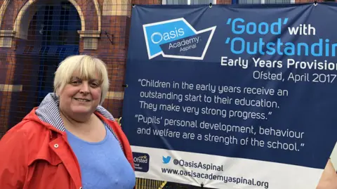 Liberal Democrats Jackie Pearcey has a red jacket, sky blue sweater and bobbed blonde hair. She is standing outside Gorton Oasis Academy.