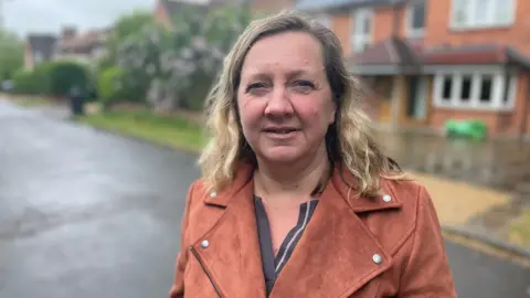 Lucy Nethsingha looking at the camera. In the background, out of focus, are some houses and hedges. She is wearing a brown suede jacket and has long blonde hair. 