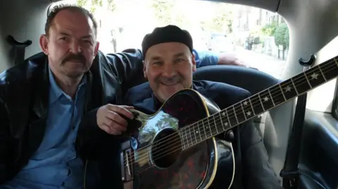 Getty Images Two people are seated in the back of a car, with one holding an acoustic guitar.