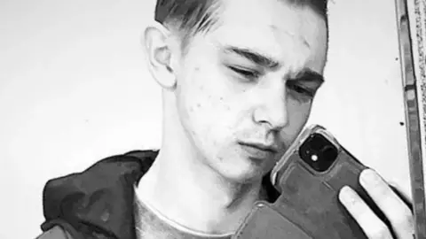 Facebook A black and white photo of Scott Jeff taking a selfie with his mobile phone. He is looking at the phone, and is wearing a dark hooded jacket. His phone has a case on it. 