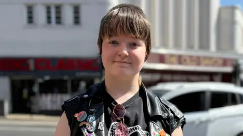 Rob Constantin/BBC Bowie is standing outside. They have a fringe and are wearing a black T-shirt and black sleeveless jacket. They are looking directly at the camera and smiling.