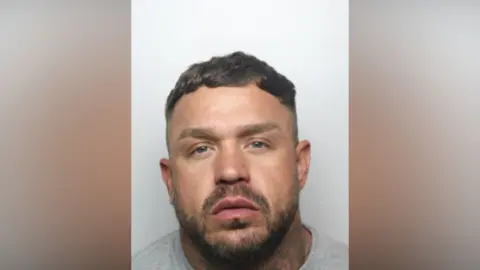 GMP Sebastian Jones has short brown hair and a beard. He wears a grey sweat shirt.