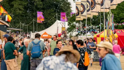 Garry Jones Photography Lots of people walk through a main 'street' in the festival, which is lined with lots of small tents and marquees. There are also brightly-patterned flags and lights strung up. The festival-goers are smiling and talking with each other, some of them with drinks in hand. 