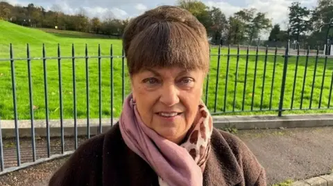 A woman, picture from the shoulders up, looking at the camera. She has brown hair, tied up and has a fringe. She is wearing a brown coat and a scarf. There is a park behind her with steel gates in front of it. 