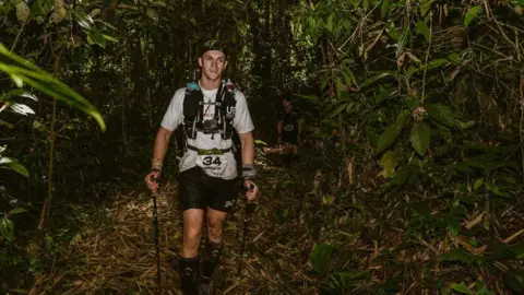 Jenny Scott Communications A man walking through a forest area. He is wearing a cap, a white t-shirt and black shorts.