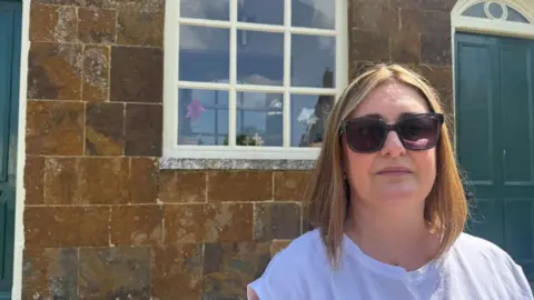 Annabel amos/bbc woman wearing sunglasses in front of a stone building with a green door.
