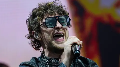 Tom Grennan wearing a black jacket and dark sunglasses, singing into a black microphone.