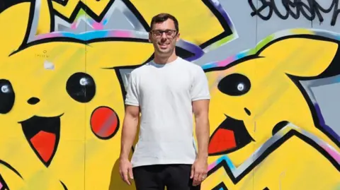 Richard Winterton Auctioneers Mr Free is pictured in a white t-shirt smiling to camera while he stands in front of a wall that has been graffitied over with images painting of the Pokémon Pikachu.