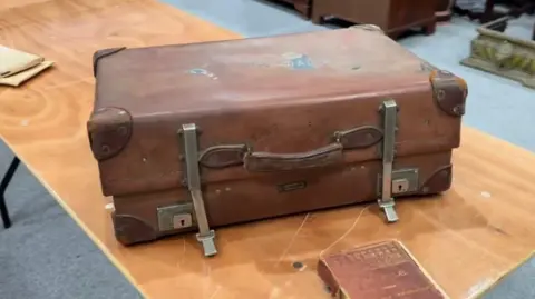 David Harper The suitcase which is brown leather with faded stickers on it. It has two locks for its metal clasps. It is sitting on a wooden table with an old, reddish coloured book in front of it.