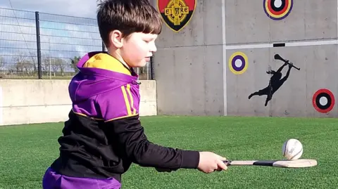 Fionn O'Neill playing hurling. The eight-year-old is balancing a sliotar on a hurling stick in a practice pitch. He has short dark hair and is wearing the black, purple and yellow sportswear of Carryduff GAC