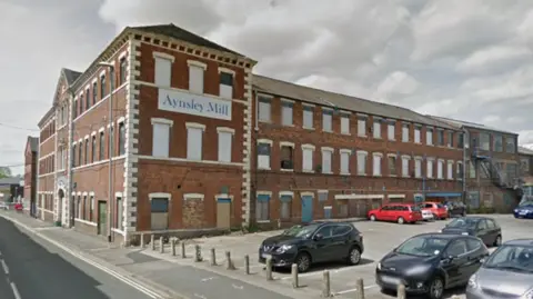 Google A three-storey mill building in red brick with white detail including the window lintels and door arches. A white sign with blue writing on the side reads "Aynsley Mill". Cars are parked in a car park in the foreground. 