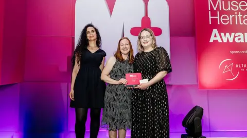 Hayley Bray Three women on the stage holding a award. They are all smiling at the camera and wearing black dresses 