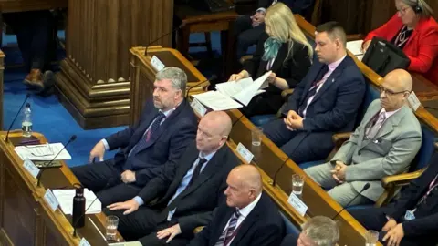 Several men in navy blue and grey suits, shirts and ties are sitting on wooden benches in a council chamber