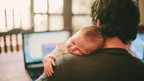 Getty Images A man holding a baby while working on a laptop. The baby is sleeping on his shoulder.