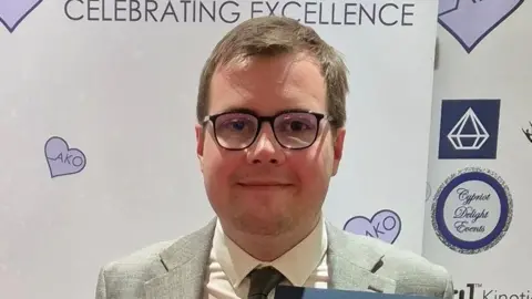 A head and shoulders image of a man, who is wearing glasses, a grey suit, white shirt and dark grey tie. He is looking straight into the camera and he has light, short, brown hair.