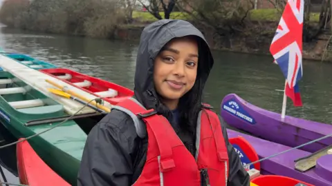 A South Asian women wearing a black waterproof jacket with a hood on. She is sitting in a bell boat wearing a red life jacket and smiling to the camera. There is a Union Jack flag on a purple boat on her right.