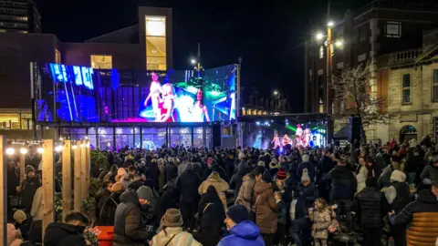 Sunderland City Council Hundreds of people gathered in Keel Square where entertainers are performing on a stage. A large screen showing the action is positioned above.
