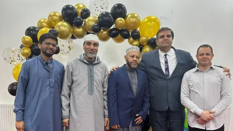 Ben Pickering Five men stand smiling in front of a gold and black balloon arch at Elaf Mosque.
