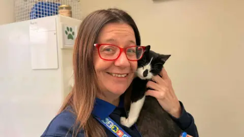 A woman with long brown hair and red framed glasses cuddles a black and white kitten on her shoulder. The woman is smiling and the kitten looks content, with its eyes closed.
