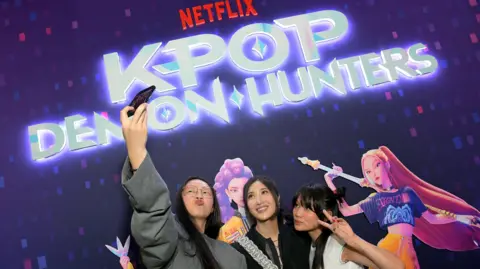 Getty Images Audrey Nuna, EJAE and Rei Ami attend the KPop Demon Hunters Special Screening at Netflix Tudum Theatre. The woman on the left is holding a camera up in the air for a selfie while the others look up and pose. Behind them is the film's logo and pictures of the animated characters which have long pink and purple hair.