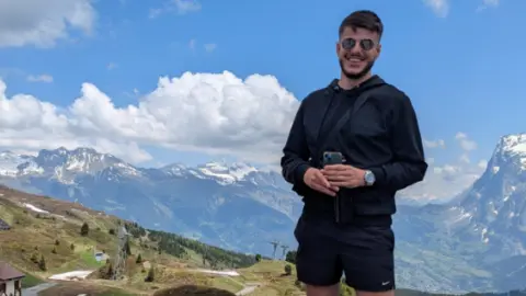 Drew Perham is pictured standing on a mountain path with a wide alpine landscape stretching out behind them. He is wearing a black jacket, black shorts, trainers and white socks, and is holding a phone in his hands. The ground around them is a mix of grass, patches of remaining snow and gravel.