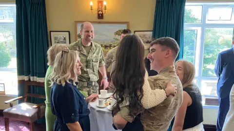 People stood around a white table smiling with cups of tea in front of them. Prince William is among them and his smiling at a man who is holding his young daughter, who has long dark brown hair