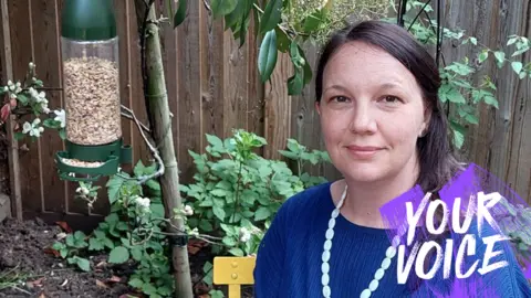 Tony Joliffe A woman with long brunette hair, wearing a blue jumper and a bead necklace stands in a garden next to a bird feeder. 