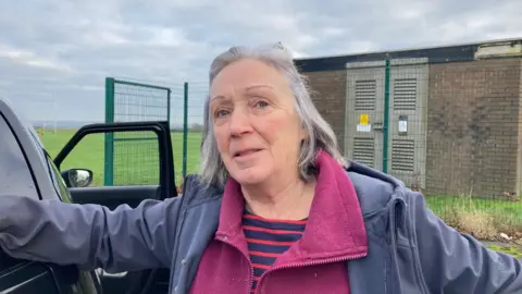 A silver-haired woman wearing a grey hooded top and plum-coloured zipped fleece and red and blue striped T-shirt underneath learning against a car. 