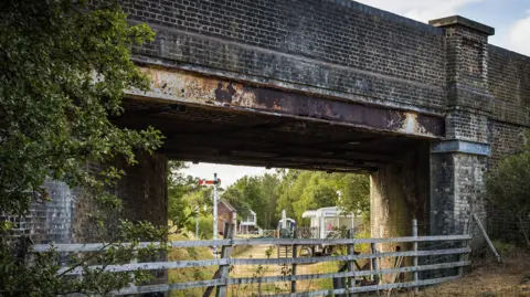 A brick railway bridge spanning a narrow passageway, with weathered steel beams visible beneath the structure. A wooden fence runs across the foreground, separating the viewer from the area under the bridge. Through the opening, several small buildings, a level-crossing gate, and a traditional railway semaphore signal can be seen in the distance. Trees, shrubs, and dry grass surround the scene.