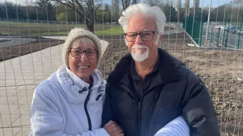 An older couple standing together outdoors in front of metal construction fencing, with upturned brown soil and a park visible behind them. The woman on the left wears a white padded jacket and glasses, and the man on the right wears a dark jacket and glasses and has white hair and a white beard. Both are smiling.