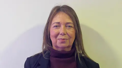 Kerrie Carmichael has long brown hair, past her shoulders, and wears a dark blue jacket over a mauve jumper, in front of a white wall.