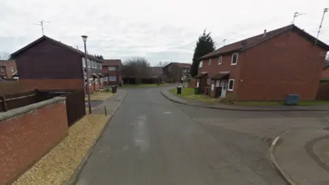 Google A residential street that splits off into side streets, with red brick houses on both sides that look like they were built in the 1980s or 1990s.