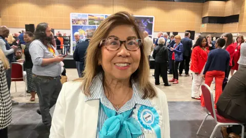 Amy Holmes/BBC A woman wearing glasses and a light jacket with a turquoise scarf and a blue “Reform UK” rosette stands in a busy indoor hall. Behind, groups of people gather and talk near a stage area, with posters, chairs, and bright overhead lighting visible.