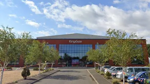 A hall-sized building with a glass frontage and KingsGate written in silver on the front. There are trees and cars parked outside it.