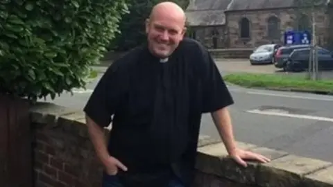 St Michael's Church, Flixton Rev Huw Thomas in clerical attire leaning on a wall near his church.