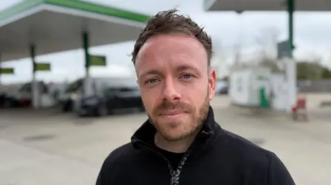 A head and shoulders image of Tom Highland. He is wearing a black top and standing on a petrol station forecourt. He is looking directly into the camera.