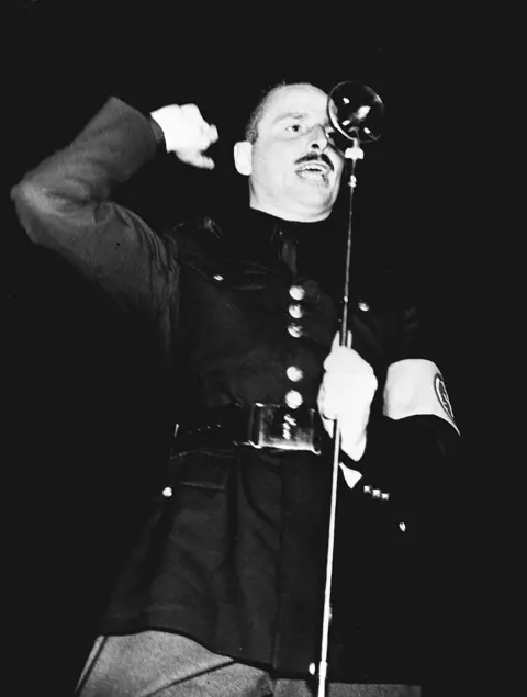 PA News A black and white photograph shows a moustached man shouting into an old-fashioned microphone. He is wearing a black military-style jacket with a belt around the waist and an arm band on the left sleeve.