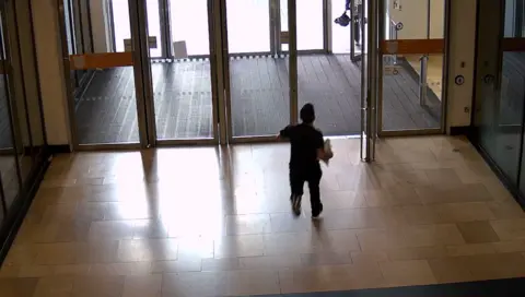 The back of a man running through big glass doors of a shopping centre. He's wearing a black top, trousers and trainers and holding a white shopping bag in his right arm. 
