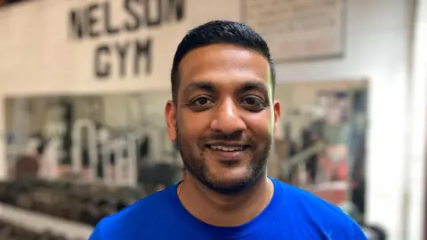 A man smiles toward the camera - he is wearing a blue T-shirt, behind him are dumbbells on a weights rack in a gym.